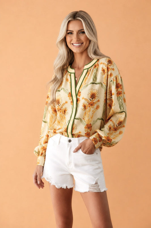 A woman modeling a long-sleeve button-up blouse with a yellow and orange floral print and green trim, paired with white distressed shorts, against a solid peach studio background.