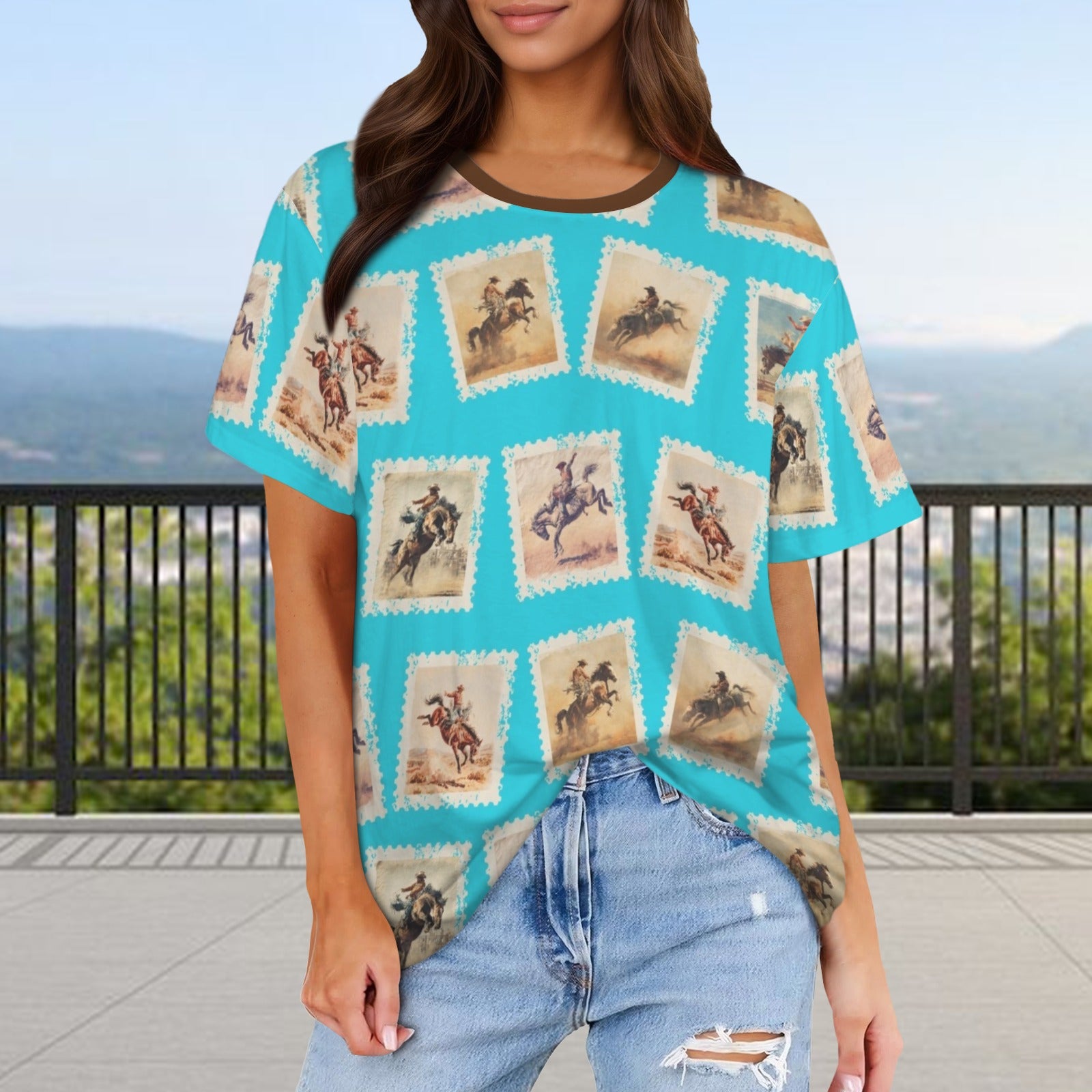 A woman wearing a turquoise short-sleeve t-shirt with a vintage stamp-like western rider pattern, paired with light-wash distressed jeans, set against an outdoor scenic background.