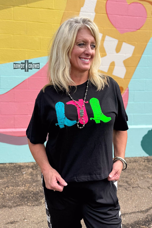 A smiling blonde woman wearing a black graphic t-shirt with colorful cowboy boot designs, standing in front of a brightly painted mural wall.