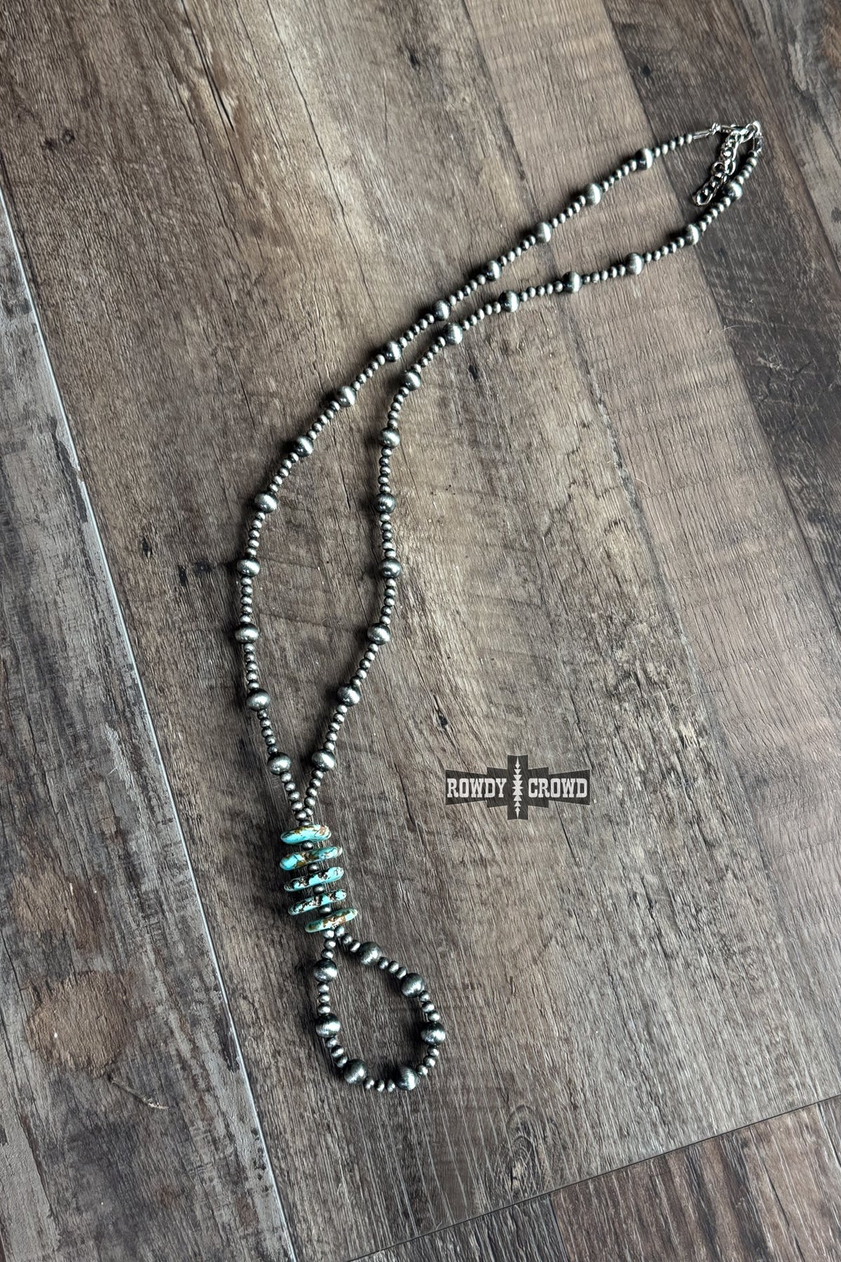 A beaded necklace with dark gray and silver beads, featuring a central cluster of turquoise-colored spacer beads and a large oval bead at the bottom, laid out on a rustic wooden floor. A small 'BOWTY CROWN' brand label is placed near the center.