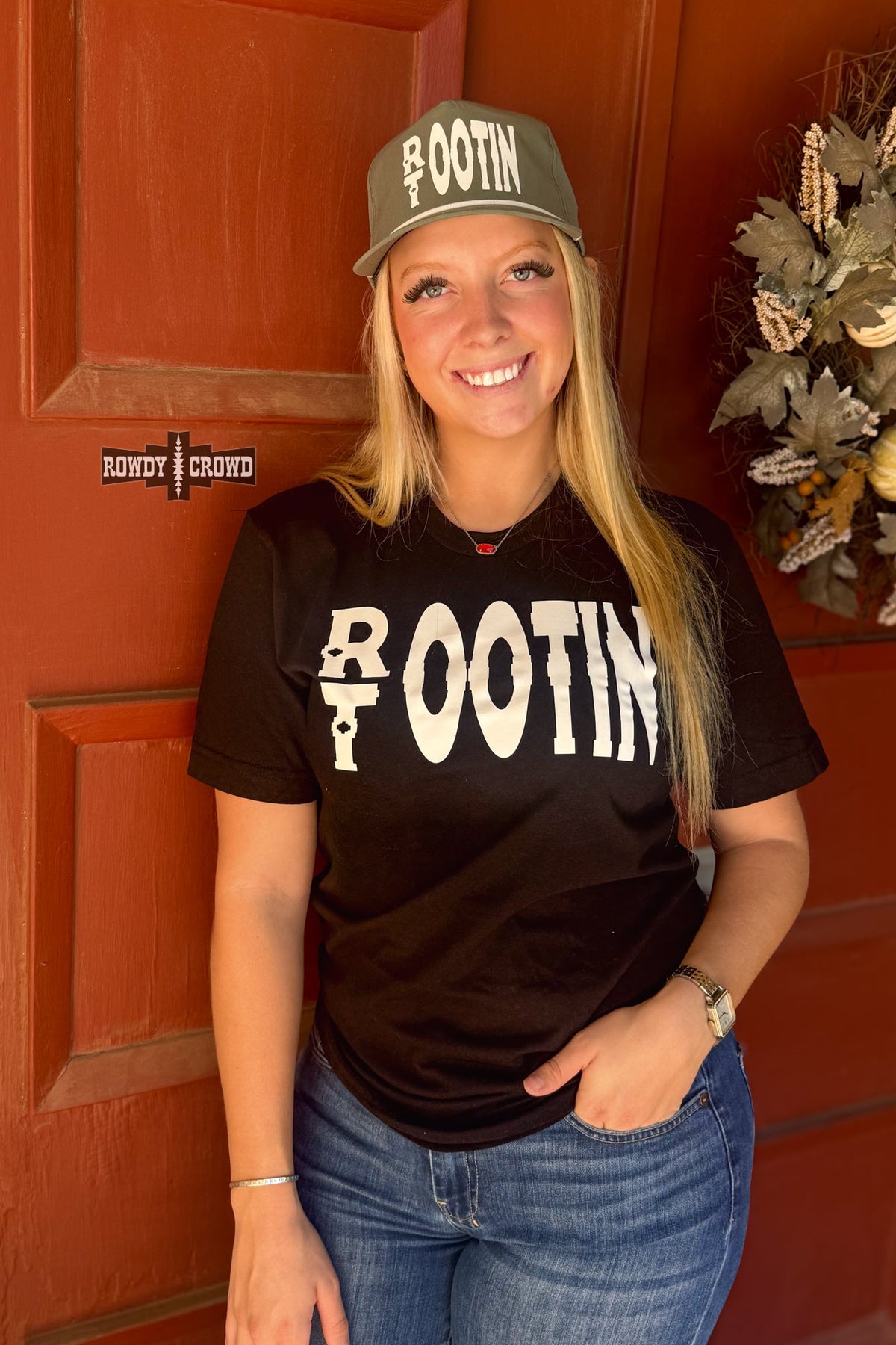 A smiling blonde woman stands in front of a reddish-brown wooden door wearing a black graphic T-shirt with large white 'ROOTIN'' lettering, blue jeans, and an olive green cap with white text. A small 'BOWOT CROWD' logo watermark appears on the left side.