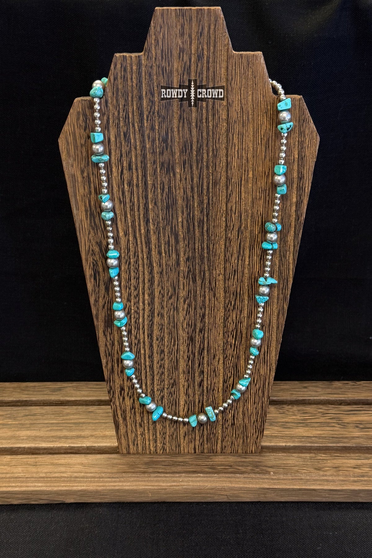 A turquoise and silver beaded necklace displayed on a dark wood stand against a black background.
