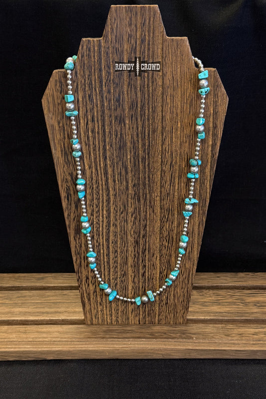 A turquoise and silver beaded necklace displayed on a dark wood stand against a black background.