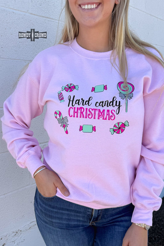 A woman wearing a light pink crewneck sweatshirt with a 'Hard candy CHRISTMAS' graphic featuring candy illustrations, paired with blue jeans, standing against a white brick wall.