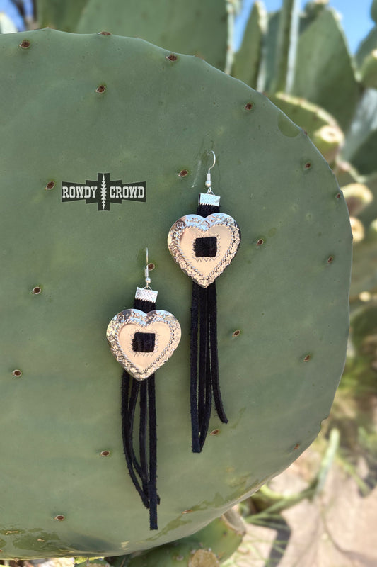 A pair of heart-shaped dangling earrings displayed on a large green cactus pad outdoors. Each earring has a light beige heart with a black square center, a silver-toned textured border, a small silver spacer, and long black fringe tassels.