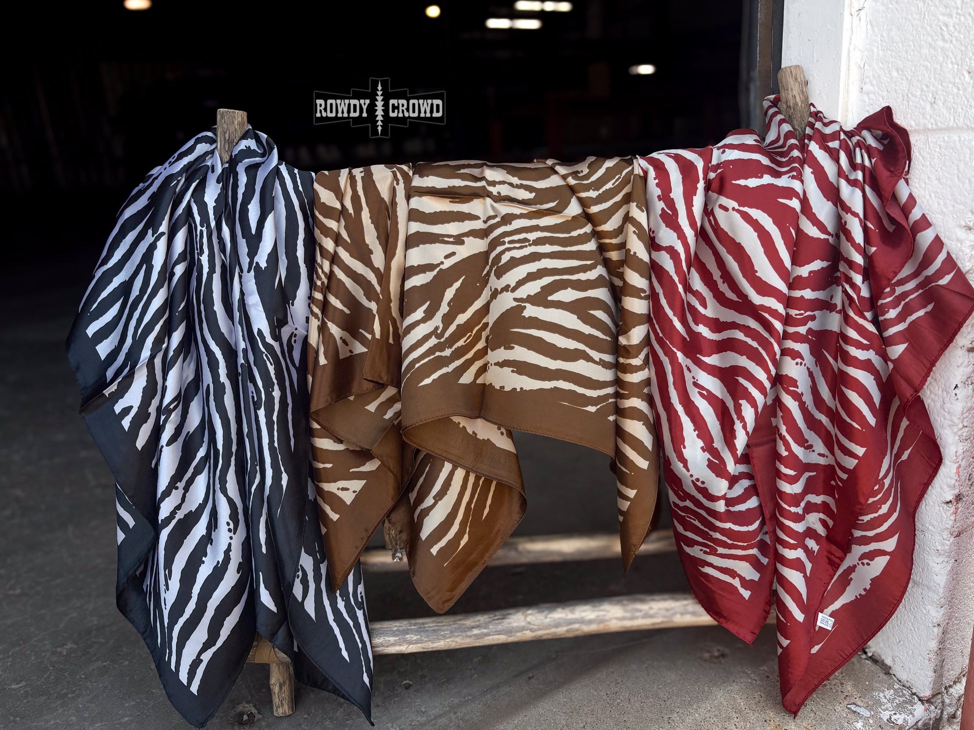 Three patterned scarves hang from wooden clips on a rustic wooden stand in a dim indoor setting. The scarves have zebra-like animal prints in black and white, brown and beige, and red and white.