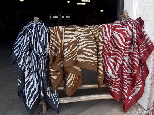Three patterned scarves hang from wooden clips on a rustic wooden stand in a dim indoor setting. The scarves have zebra-like animal prints in black and white, brown and beige, and red and white.