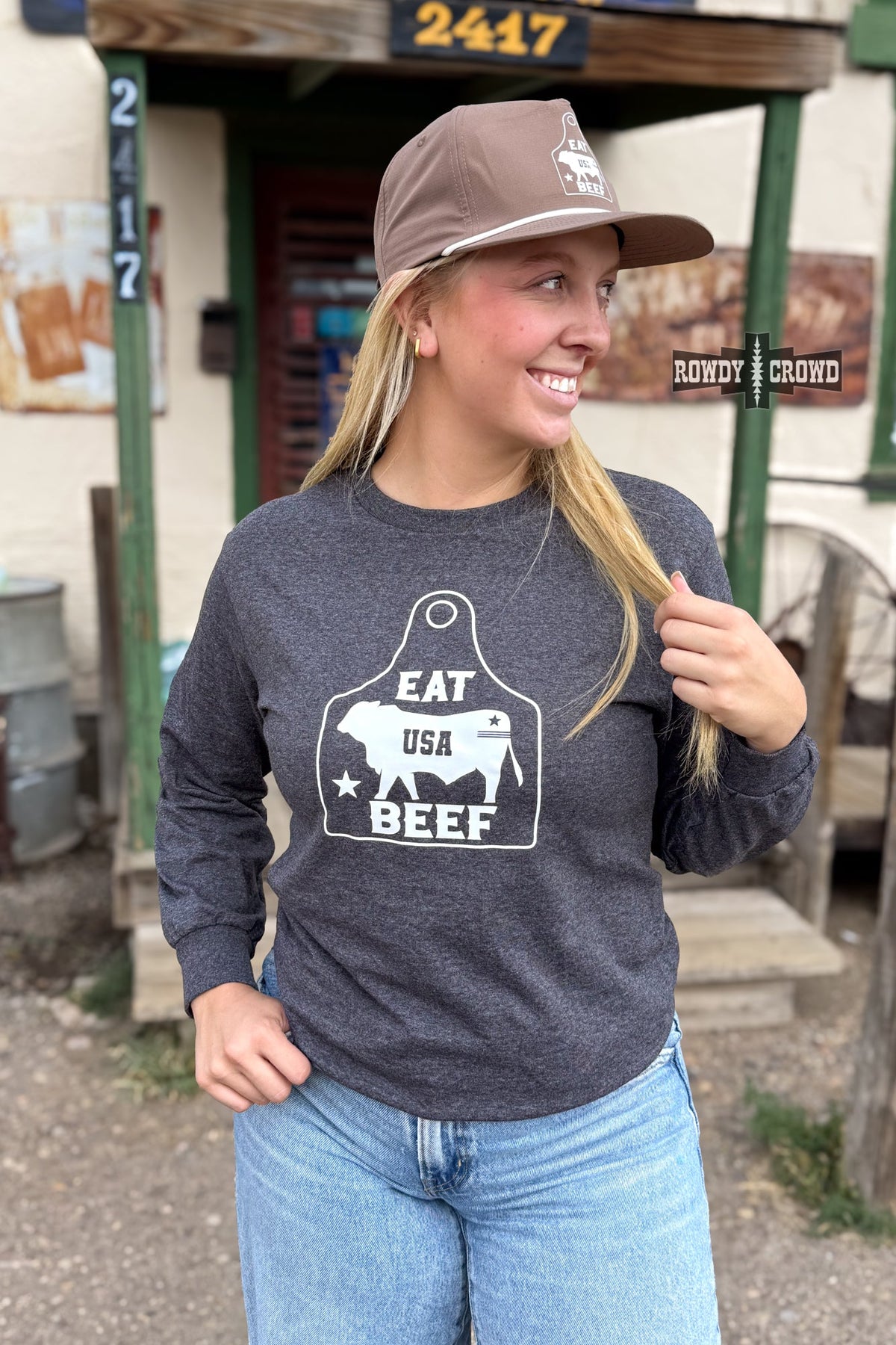 A woman wearing a dark heather gray long-sleeve t-shirt with a white graphic that reads 'EAT USA BEEF' inside a cow-shaped outline, paired with a brown baseball cap and blue jeans, standing in front of a rustic western-style building.