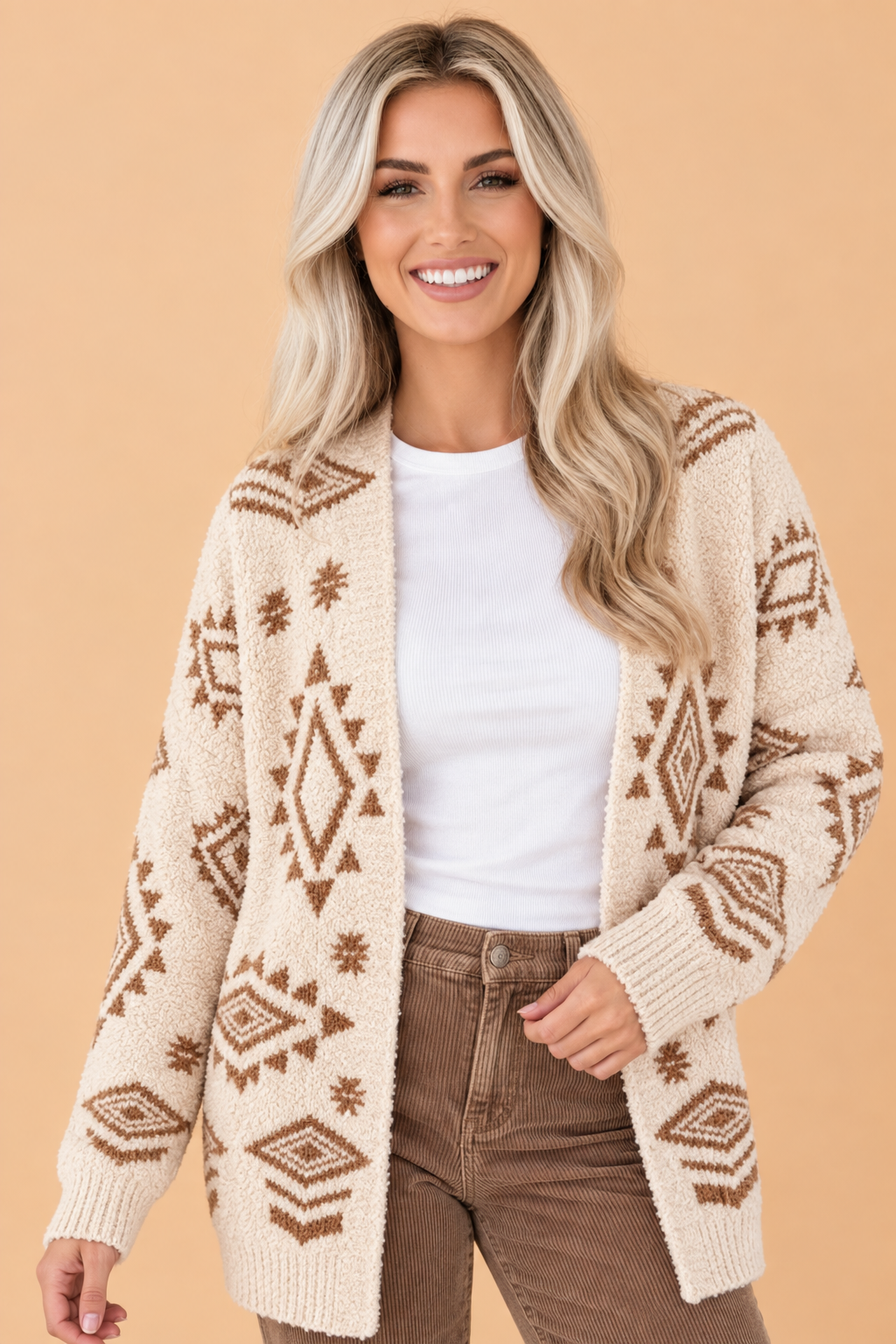 A smiling blonde woman wearing a cream knit cardigan with brown Southwestern geometric patterns, a white t-shirt, and brown pants, set against a solid beige background.