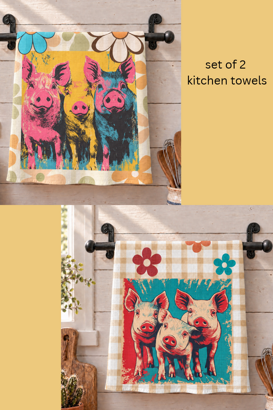 Set of two kitchen towels hanging on black metal racks against a light wood wall. The top towel has a colorful pop-art style print of three pigs in pink, yellow, blue, and black, with a cream border and flower motifs.