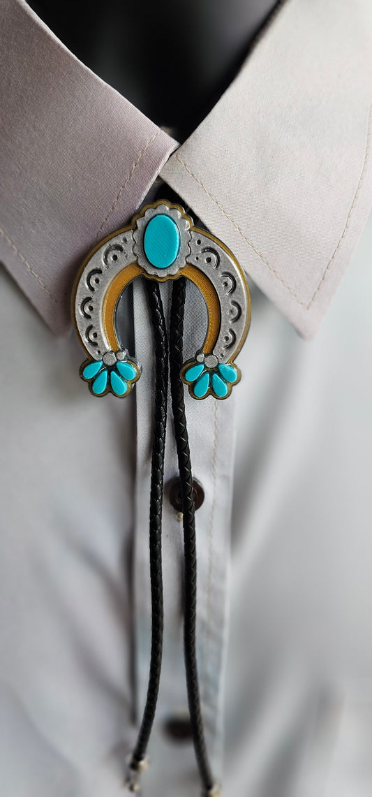 A close-up of a western-style bolo tie worn under a light gray dress shirt collar. The bolo features a silver-toned horseshoe-shaped metal frame with turquoise stones, a gold-toned inner arch, and matching turquoise flower details at the ends, with black cord ties hanging below.